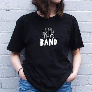 I'm With The Band® logo T-shirt with white print on black T being worn by a faceless woman standing against a white painted brick wall