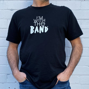 I'm With The Band® logo T-shirt with white print on black T being worn by a faceless man standing against a white painted brick wall