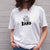 I'm With The Band® logo T-shirt with black print on white T being worn by a faceless woman standing against a white painted brick wall