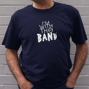 I'm With The Band® logo T-shirt with white print on navy T being worn by a faceless man standing against a white painted brick wall