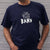 I'm With The Band® logo T-shirt with white print on navy T being worn by a faceless man standing against a white painted brick wall