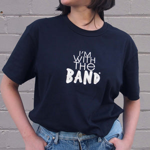 I'm With The Band® logo T-shirt with white print on navy T being worn by a faceless woman standing against a white painted brick wall