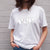 I'm With The Band® Bubble Pop T-shirt with iridescent white print on white T being worn by a faceless woman standing against a white painted brick wall