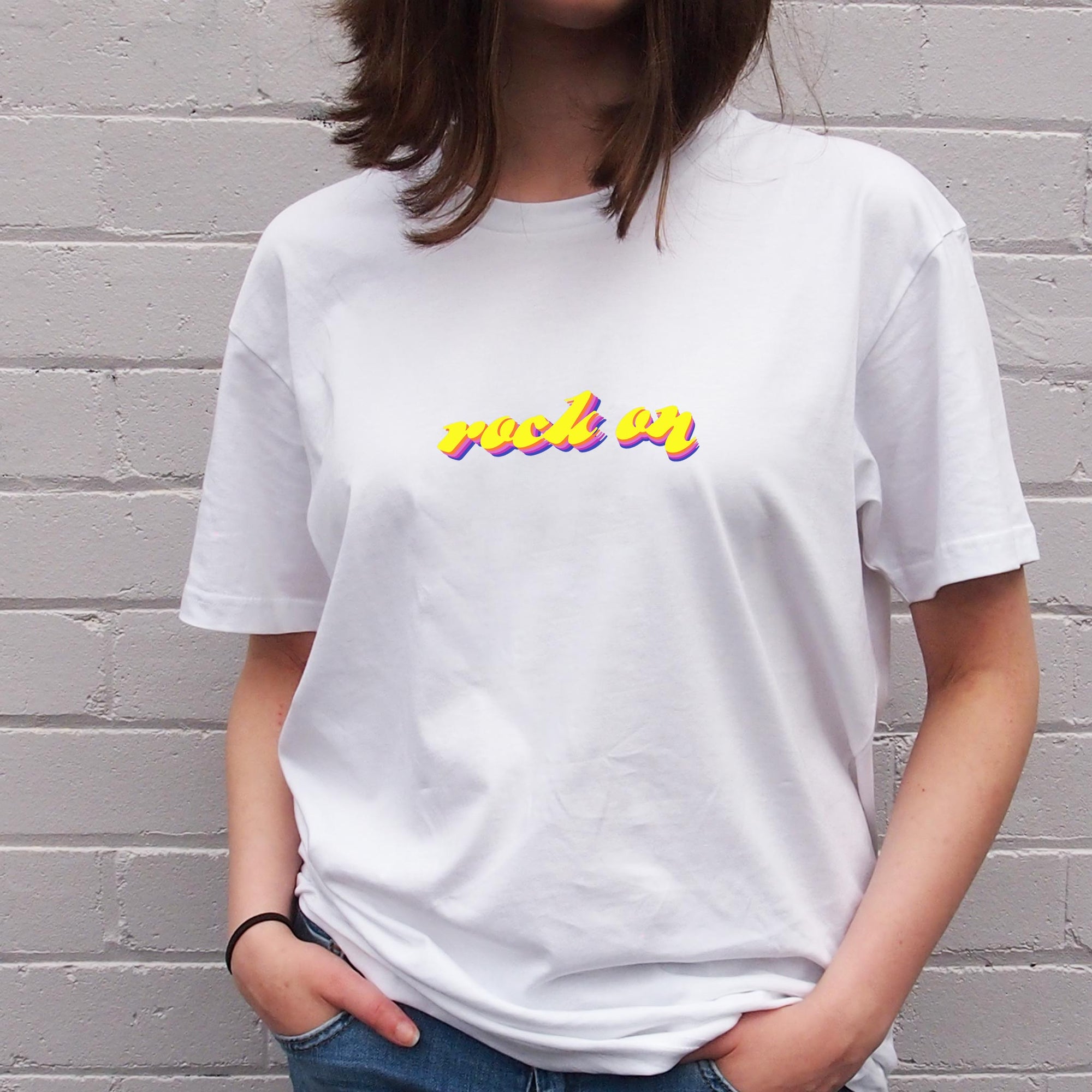 I'm With The Band® Rock On T-shirt with bright multi-colour print on white T being worn by a faceless woman standing against a white painted brick wall