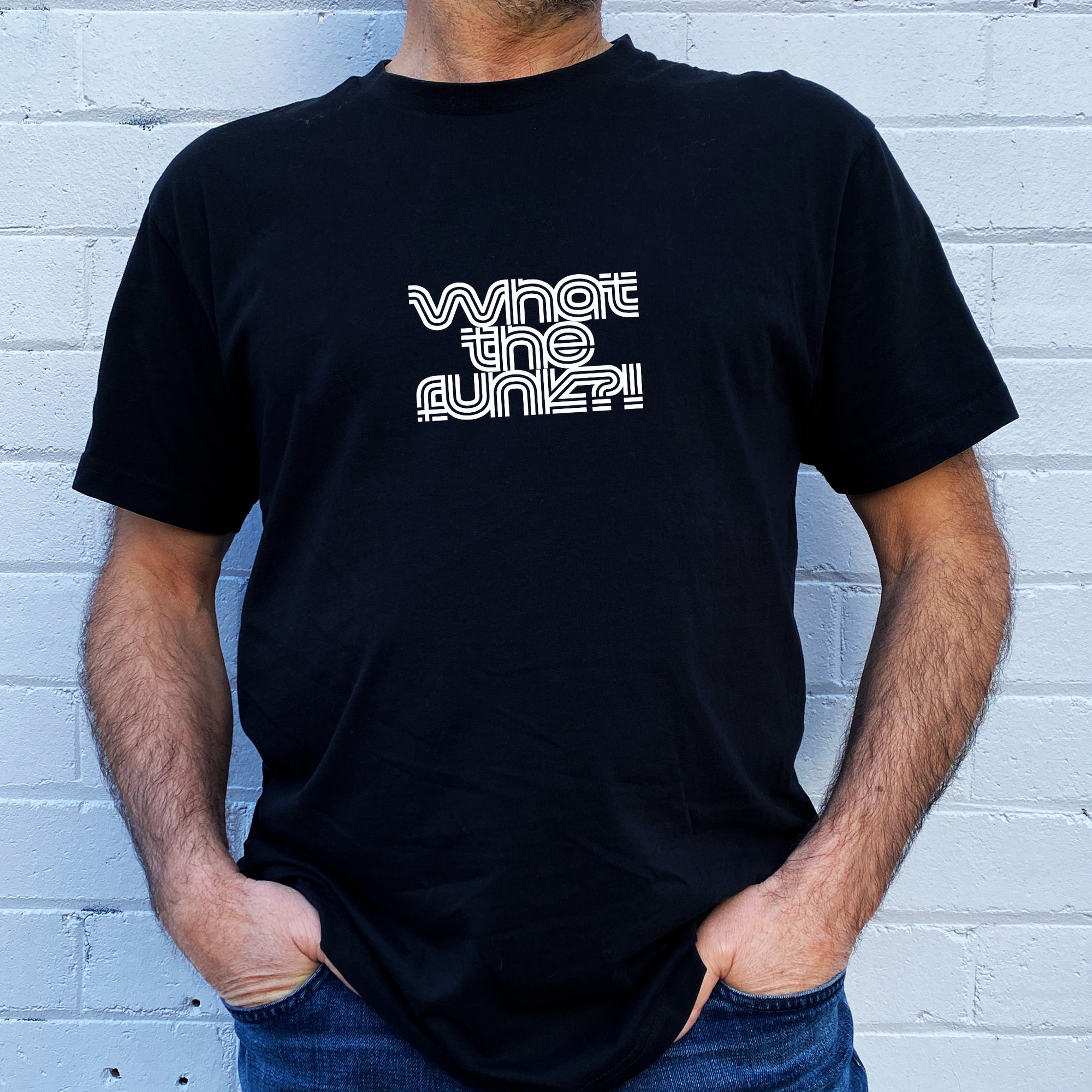 I'm With The Band® What The Funk? T-shirt with white print on black T being worn by a faceless man standing against a white painted brick wall