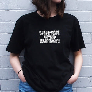 I'm With The Band® What The Funk? T-shirt with white print on black T being worn by a faceless woman standing against a white painted brick wall