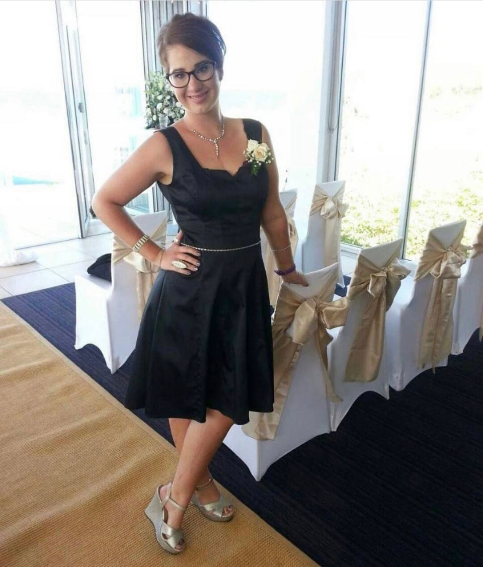 Emma is a 20-something girl. She looks happy and is dressed in her bridesmaid outfit with a corsage on her wrist.