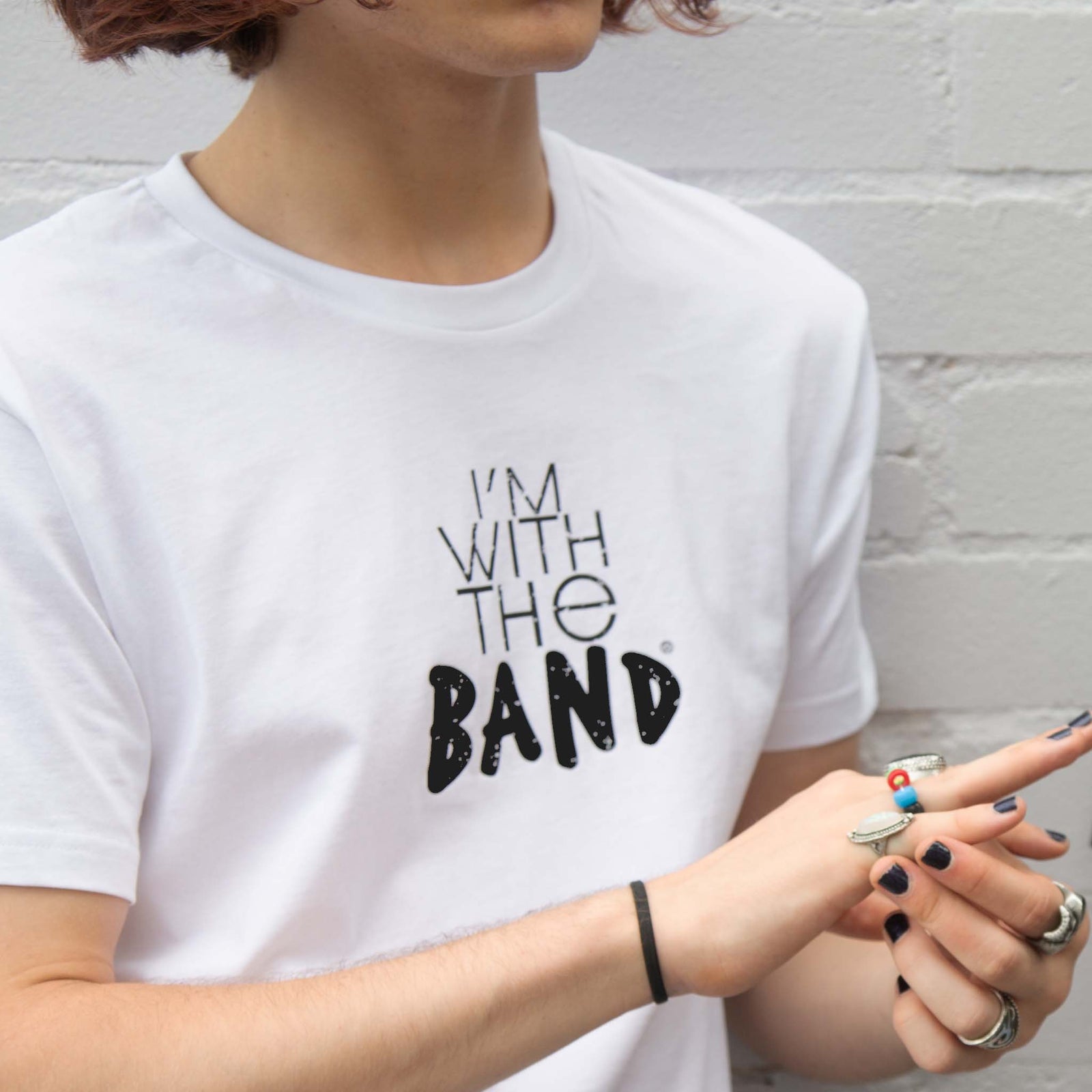 I'm With The Band® logo T-shirt with black print on white T being worn by a cool young man standing against a white painted brick wall