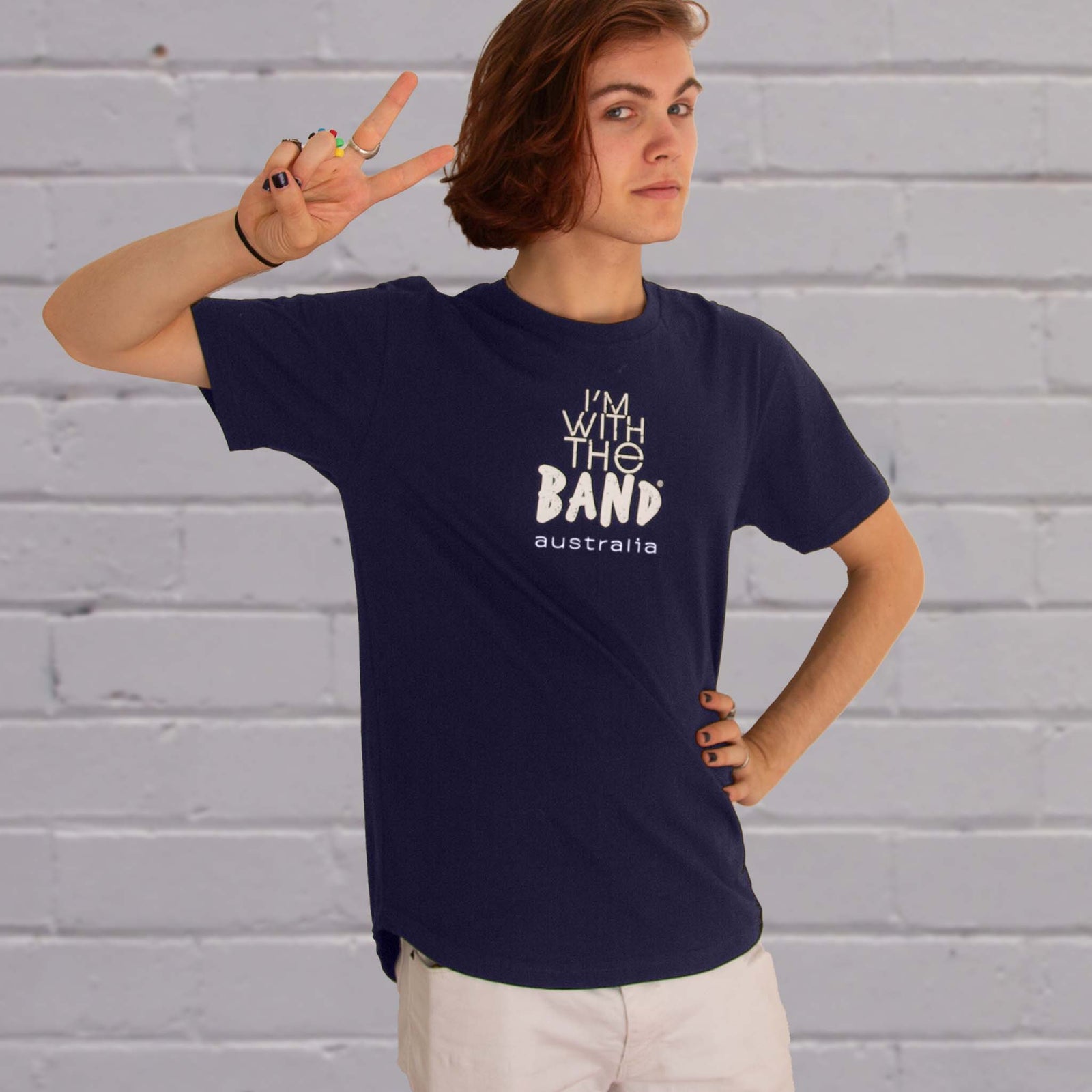 I'm With The Band® Australia logo T-shirt with white print on navy T being worn by a cool young woman standing against a white painted brick wall. He is looking into the camera and making a V sign with his fingers.