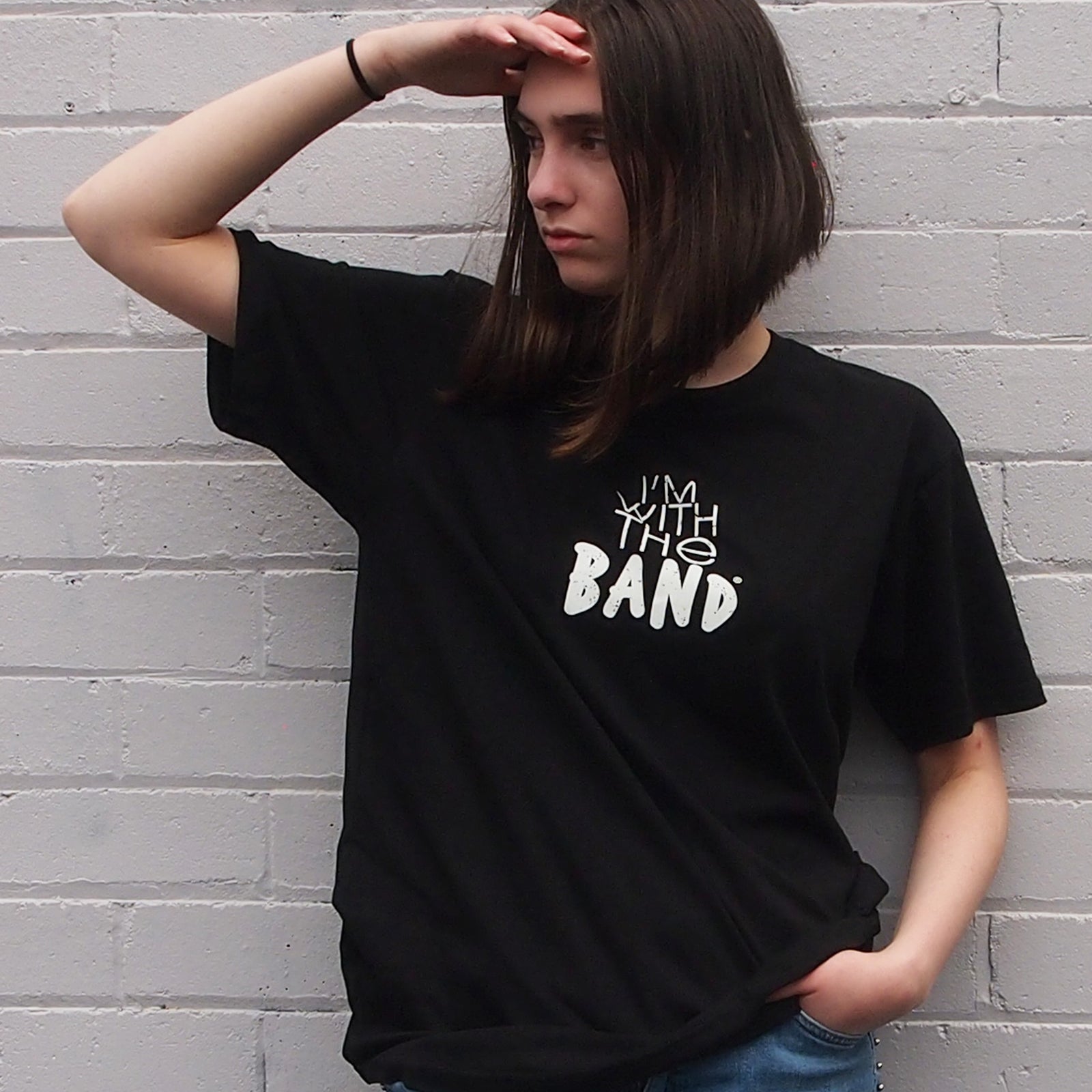 I'm With The Band® logo T-shirt with white print on black T being worn by a cool woman standing against a white painted brick wall