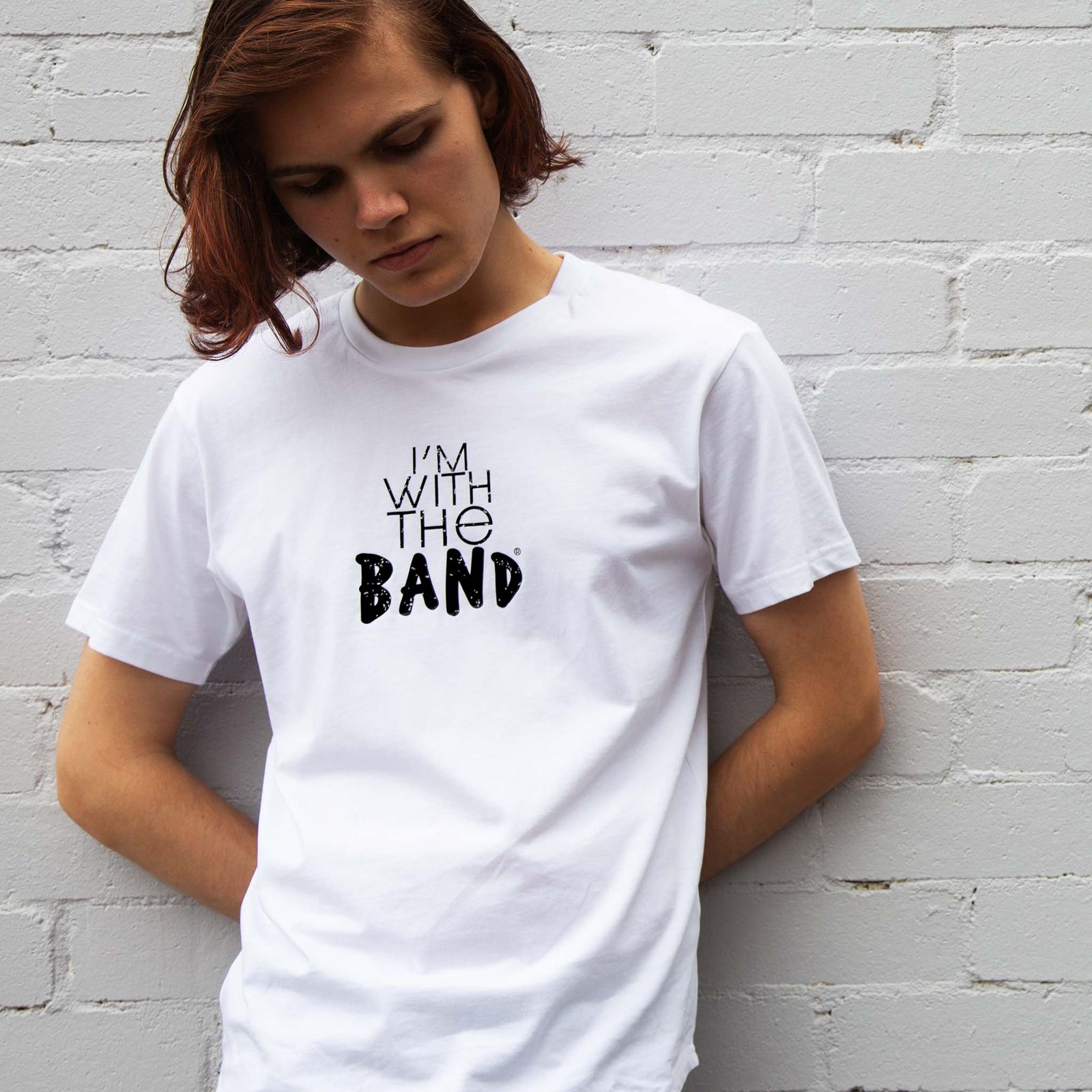 I'm With The Band® logo T-shirt with black print on white T being worn by a cool young man standing against a white painted brick wall
