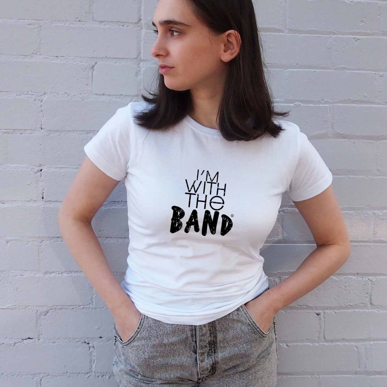 I'm With The Band® black logo print on white close-fit women's T-shirt on cool young woman who is looking down the street to her right and standing in front of a painted brick wall