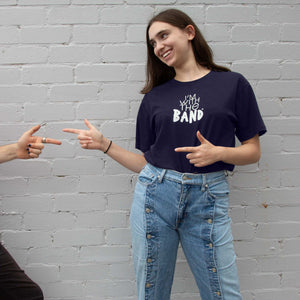 I'm With The Band® logo T-shirt with white print on navy T being worn by a smiling young woman standing against a white painted brick wall. She is playfully pointing to someone off-screen and they are pointing back at her.