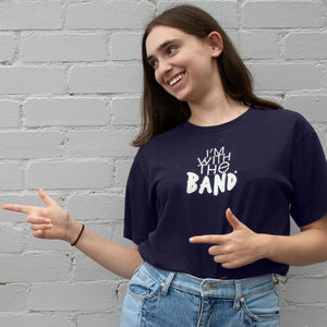 I'm With The Band® logo T-shirt with white print on navy T being worn by a smiling young woman standing against a white painted brick wall. She is playfully pointing to someone off-screen.