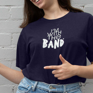 I'm With The Band® logo T-shirt with white print on navy T being worn by a faceless but smiling young woman standing against a white painted brick wall. She is playfully pointing to someone off-screen.