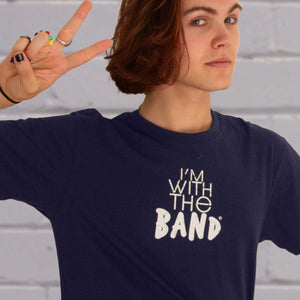I'm With The Band® logo T-shirt closeup with white print on navy T being worn by a cool young man standing against a white painted brick wall