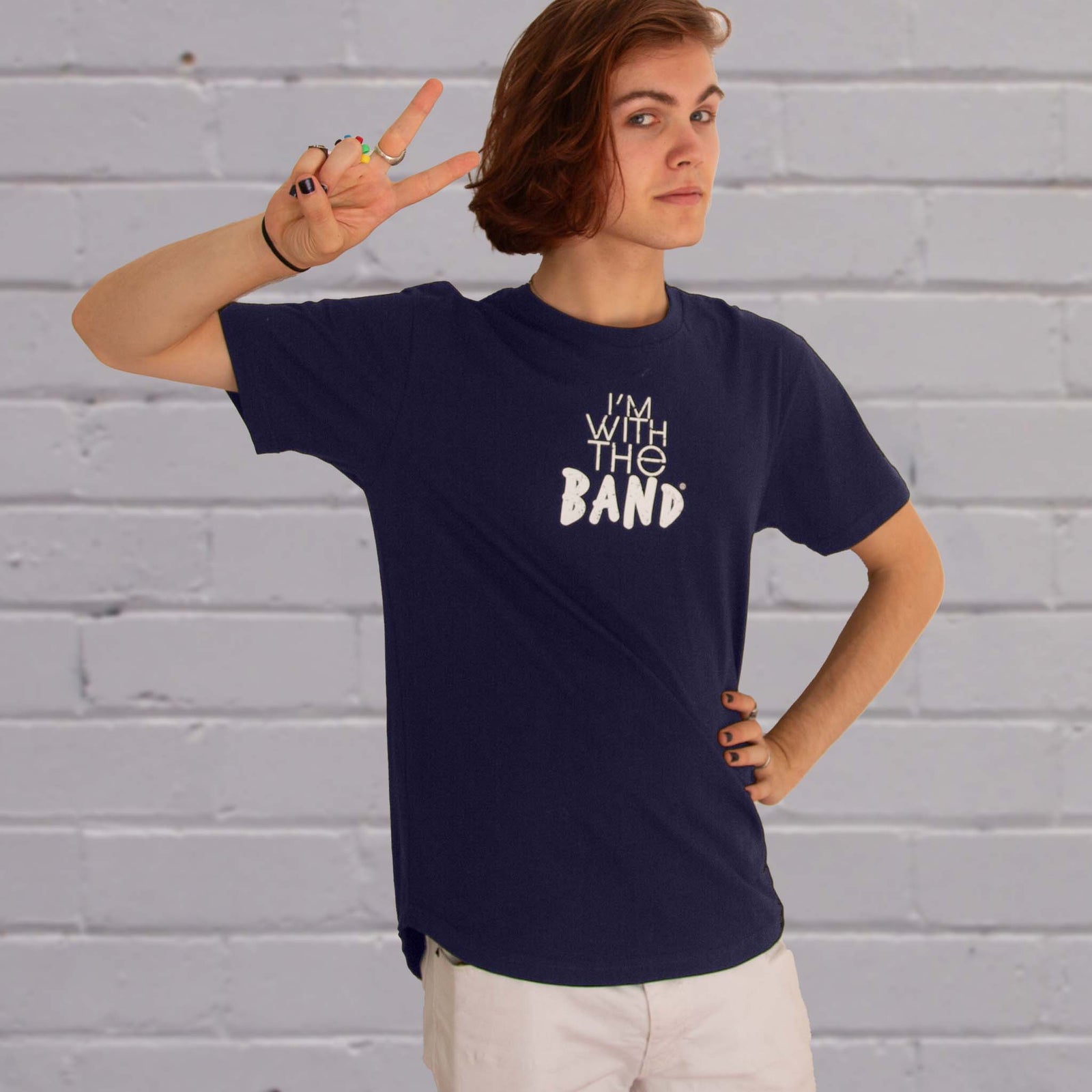 I'm With The Band® logo T-shirt closeup with white print on navy T being worn by a cool young man making a V sign with fingers and standing against a white painted brick wall